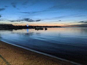 A calm waterfront with a distant illuminated tower, a sandy beach, and a calm sea.