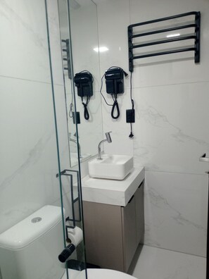 A modern bathroom with a glass shower enclosure, a white sink, and two hair dryers mounted on the wall. - Move Oásis Tambaú (Joao Pessoa)