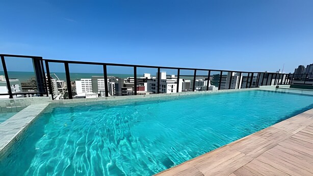 A rooftop pool with clear blue water, a wooden deck, and a view of the city skyline. - Move Oásis Tambaú (Joao Pessoa)