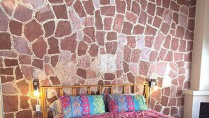 Une chambre avec un mur en pierre, un lit recouvert d’une courtepointe aux couleurs vives, une table de chevet et une lampe.