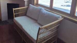 Un canapé en osier avec des coussins, dans une pièce dont un mur est en briques et qui possède une fenêtre donnant sur une cour.