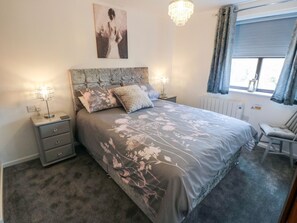 A bedroom with a bed, nightstands, a chair, a chandelier, a window with curtains, and a wall-mounted picture. - Cha Cha's Cottage (Whitby)