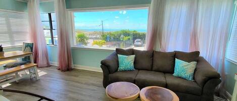 A living room with a brown sofa, two round wooden side tables, a ceiling fan, and a view of the ocean through large windows.