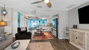 A modern living room with a sofa, coffee table, and a dining area with a table and chairs. There is a kitchen in the background with white cabinets and a black refrigerator. A ceiling fan is visible, and a TV is mounted on the wall. - Luxurious New Heated Pool & Spa! 4 mins to beach! (Seminole)