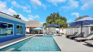 A swimming pool with lounge chairs and umbrellas, a thatched-roof structure, and a blue building. - Luxurious New Heated Pool & Spa! 4 mins to beach! (Seminole)