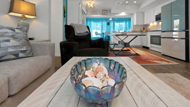 A modern living room with a sofa, a glass bowl on a wooden table, a massage chair, and a kitchen in the background. - Luxurious New Heated Pool & Spa! 4 mins to beach! (Seminole)
