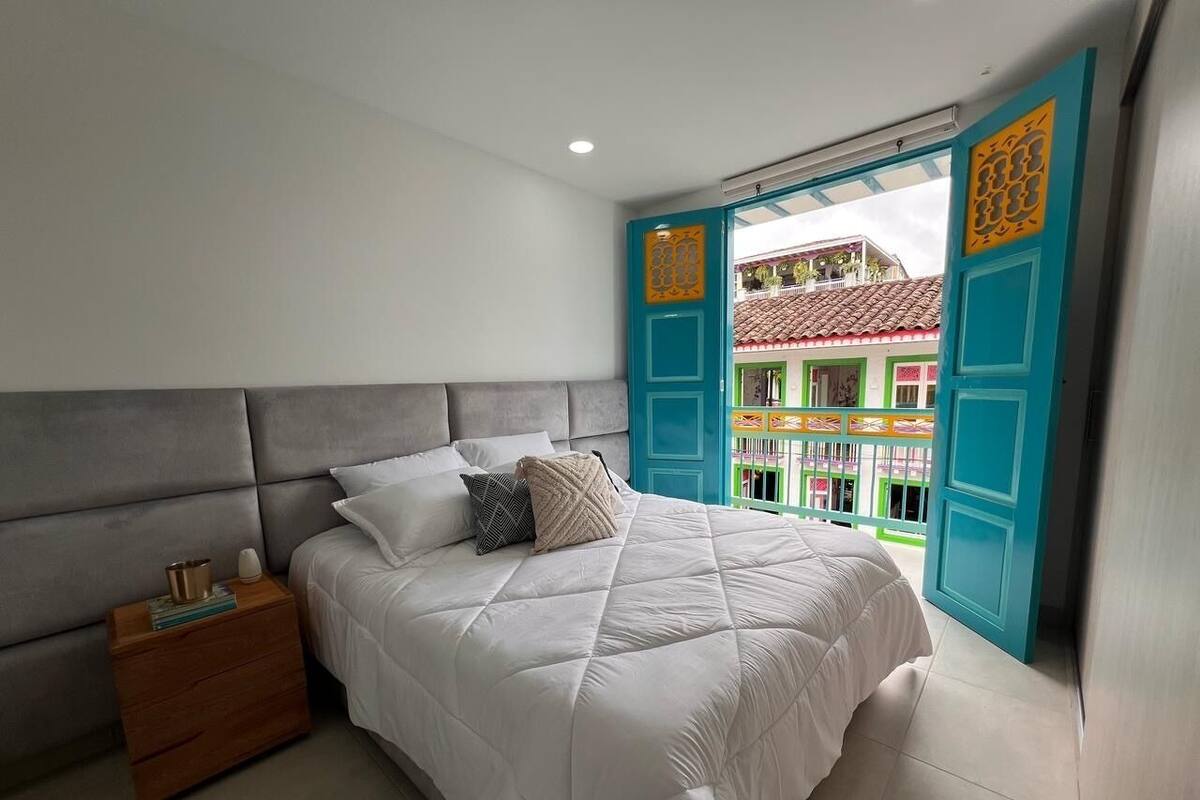 Un dormitorio con cama, mesita de noche y vistas a un edificio colorido a través de un ventanal abierto.