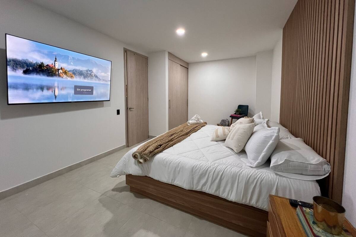 Un dormitorio moderno con una cama grande, un televisor de pantalla plana en la pared, un cabecero de madera y una mesita de noche con un libro y una taza.