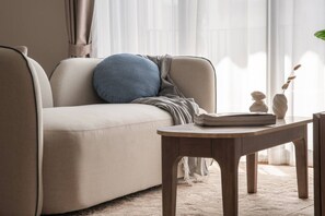 A neutral-toned sofa with a blue pillow and a grey throw blanket, a wooden side table with a book and a vase, and a white rug on a carpeted floor. - Cozy 2br Apartment Pool View- canvas- bangtao area (Choeng Thale)