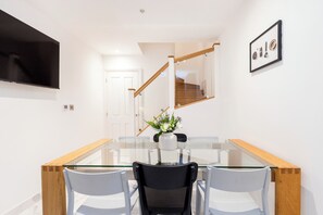 A dining area with a glass table, chairs, a vase with flowers, a TV mounted on the wall, and a staircase. - Beautiful 3 Bed House on The Edge of Hyde Park (London)