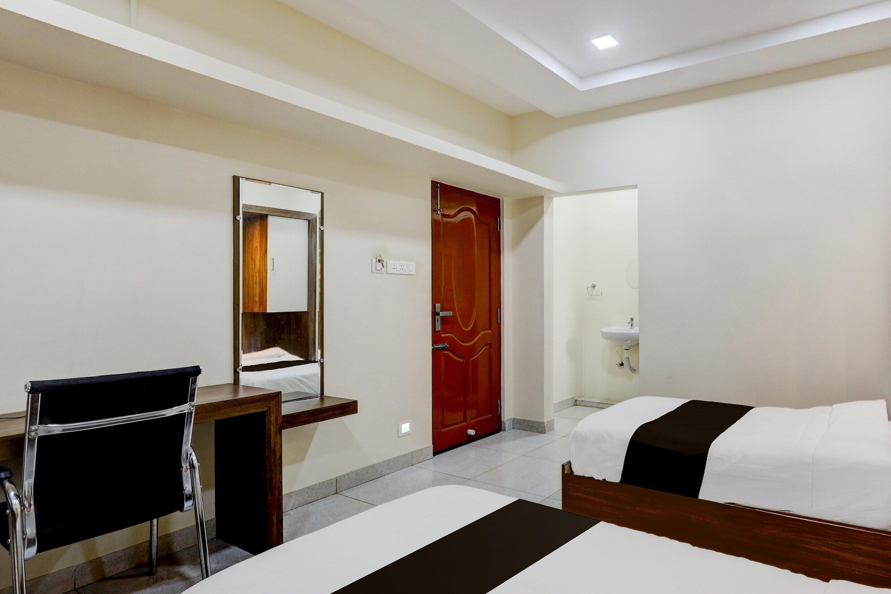 Habitación de hotel con cama, escritorio, silla, espejo, puerta y lavamanos.
