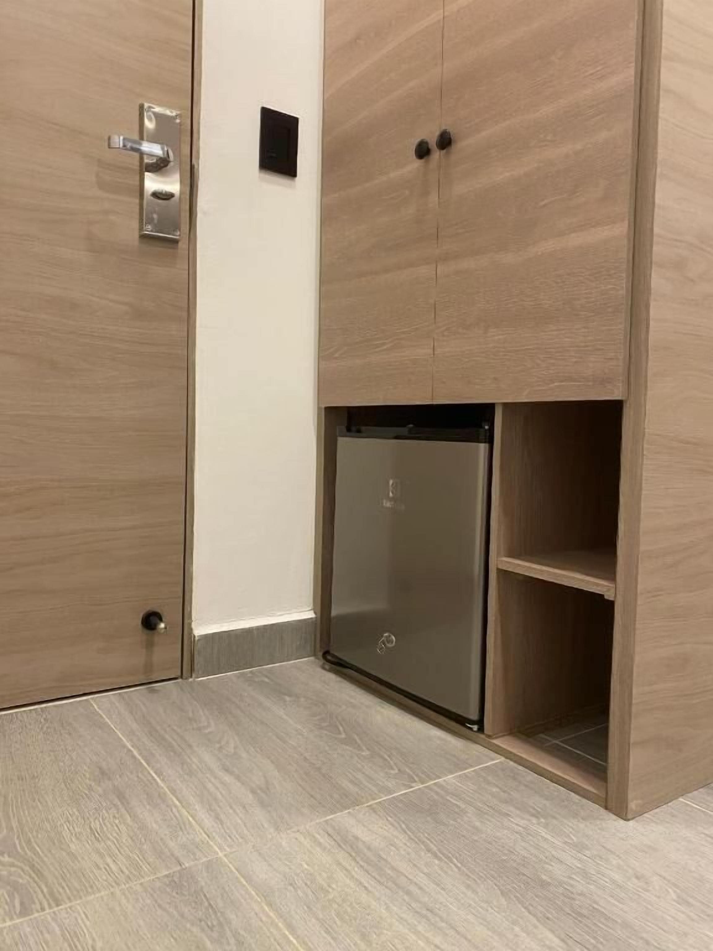 A compact room with a built-in refrigerator, a door with a handle, and a small shelf unit.