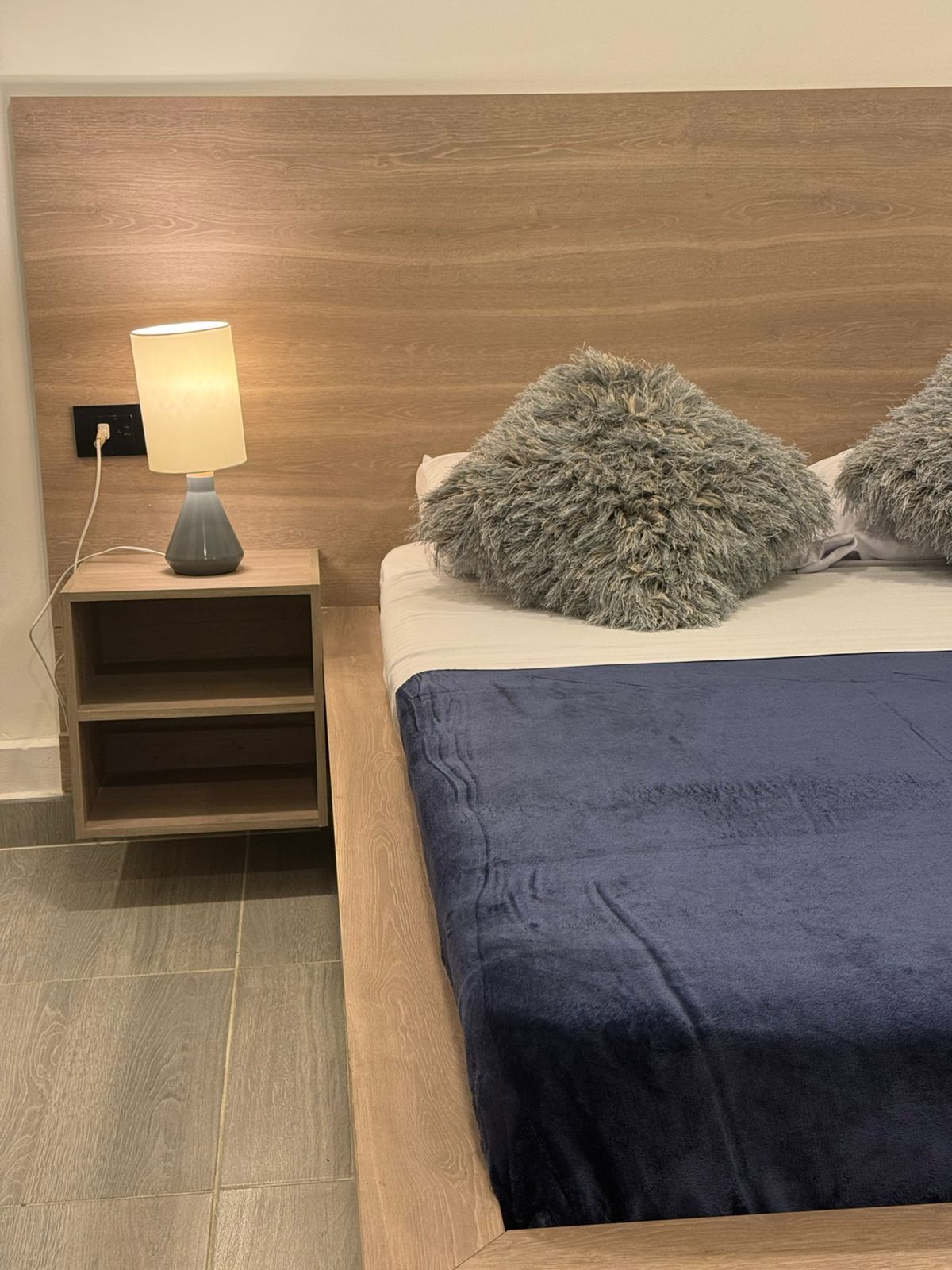 A bed with a dark blue blanket, two fluffy pillows, a bedside lamp, and a wooden nightstand.