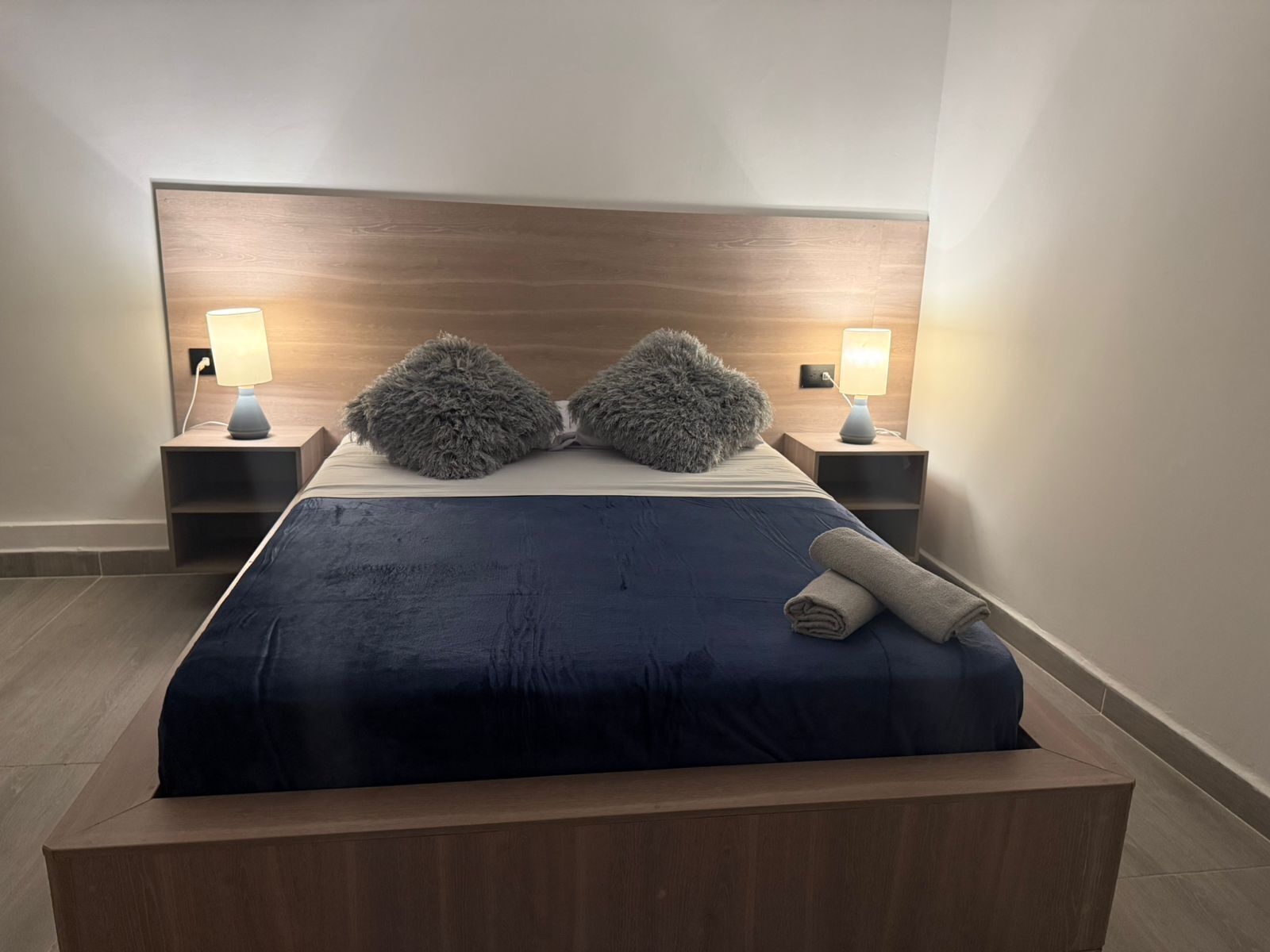 Hotellihuone, jossa on suuri sänky, kaksi yöpöytää valaisimineen, sängynpääty ja sängylle asetettu rullattu pyyhe.
