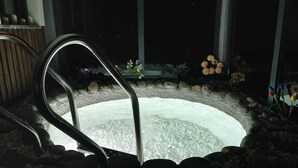 Un bain à remous entouré de rochers et de plantes, avec un banc en bois et une échelle en métal.