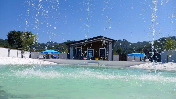 Une piscine avec des jets d’eau, un espace de repos couvert et une vue sur des collines en arrière-plan.
