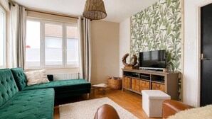 A living room with a green sofa, a white ottoman, a television on a wooden stand, and a large window with curtains.