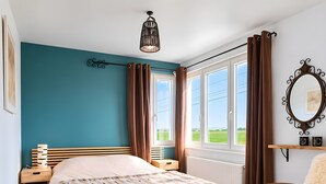 A bedroom with a bed, a nightstand, a chair, a window with curtains, a hanging light fixture, and a mirror on the wall.