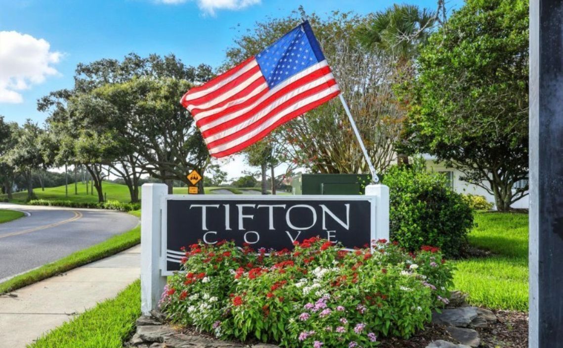 Un quartier résidentiel avec un panneau indiquant « Tifton Cove », sur lequel on peut voir un drapeau, une route et de la végétation.