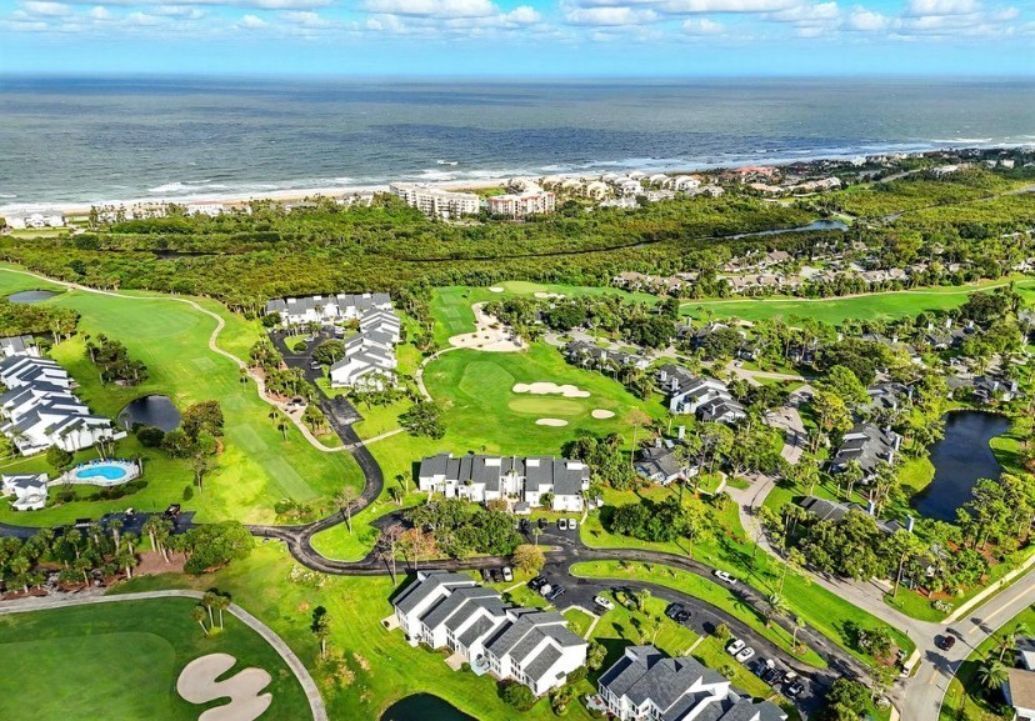 An aerial view of a golf course with residential buildings, a swimming pool, and a view of the ocean.