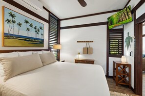 A bedroom with a large bed, a painting of palm trees, a television, and a wooden nightstand. - Kiahuna #44 • A/C • REMODELED & UPDATED Ground-Floor End Unit • 2 Min to Beach (Koloa)