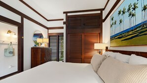A hotel room with a large bed, wooden headboard, a painting of palm trees, a wooden wardrobe, and a desk with a lamp. - Kiahuna #44 • A/C • REMODELED & UPDATED Ground-Floor End Unit • 2 Min to Beach (Koloa)