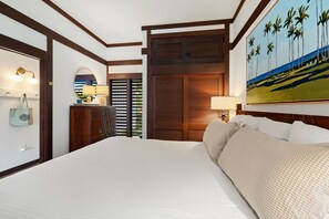 A hotel room with a large bed, wooden headboard, a painting of palm trees, a wooden wardrobe, and a desk with a lamp. - Kiahuna #44 • A/C • REMODELED & UPDATED Ground-Floor End Unit • 2 Min to Beach (Koloa)