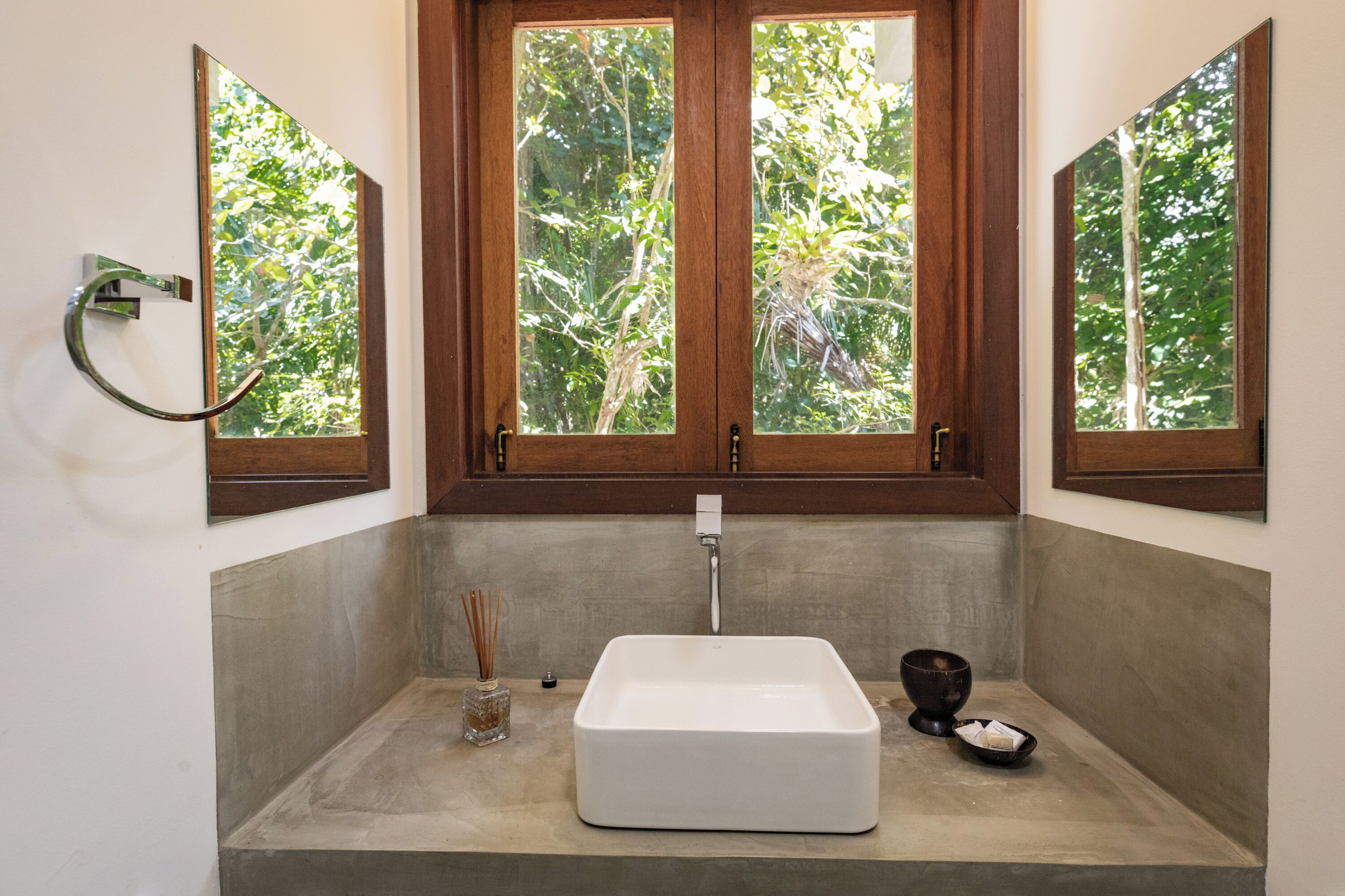 Une salle de bain avec un lavabo blanc, un cadre de fenêtre en bois et une vue sur la verdure à l’extérieur.