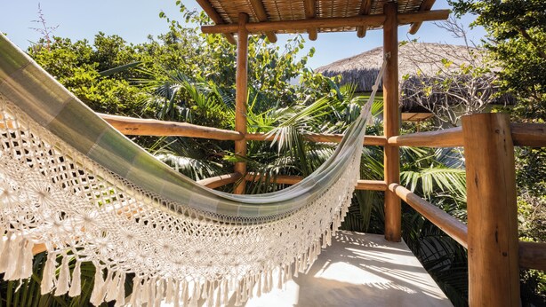 A hammock suspended between wooden beams in a lush garden with a thatched roof structure. - Pousada Colmeia (Santa Cruz Cabrália)
