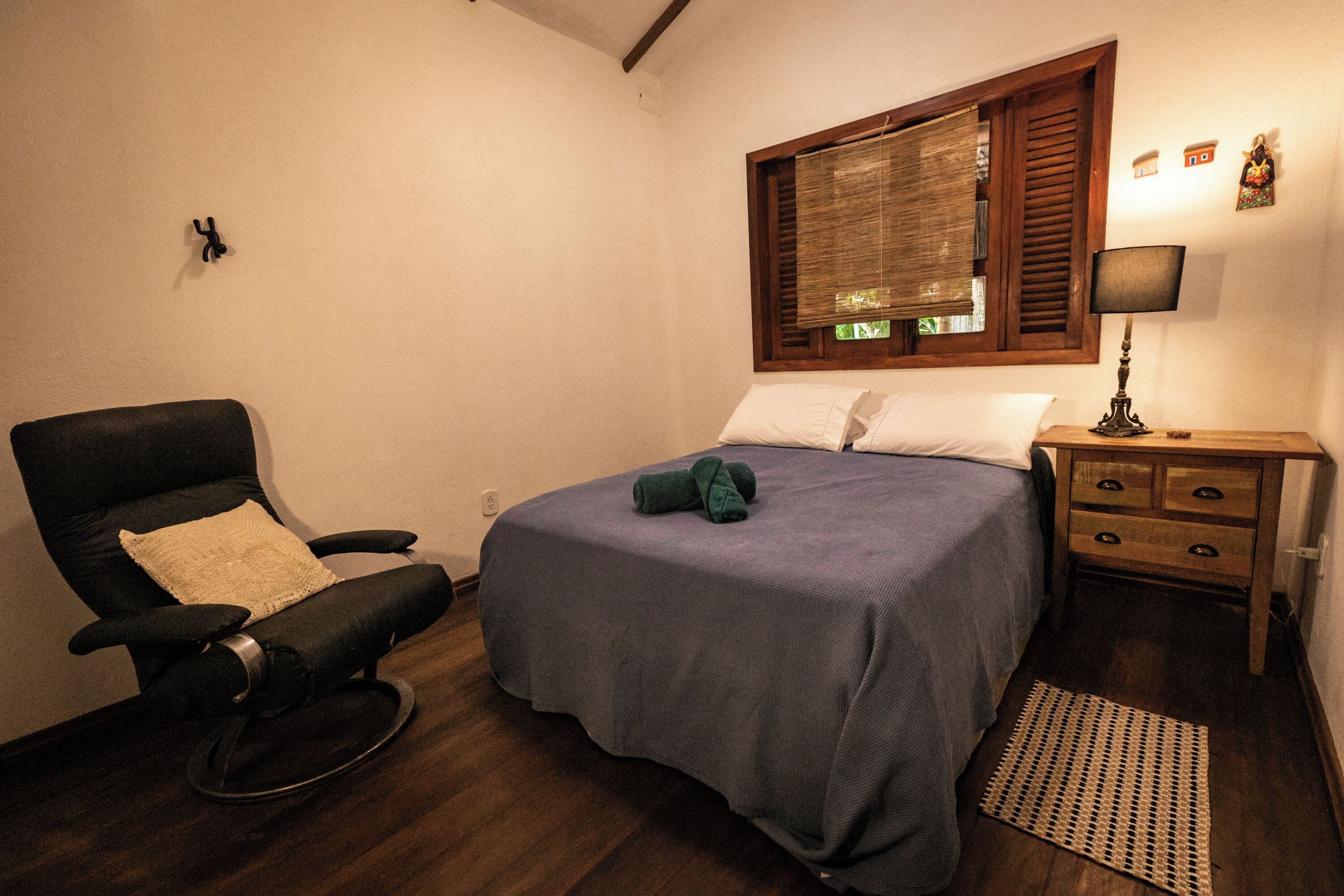 A bedroom with a bed, a rocking chair, a wooden nightstand, a lamp, and a window with blinds.
