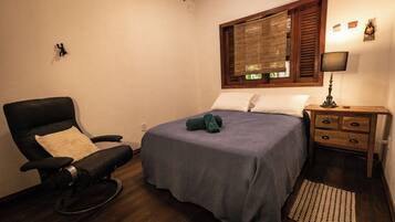 A bedroom with a bed, a rocking chair, a wooden nightstand, a lamp, and a window with blinds.