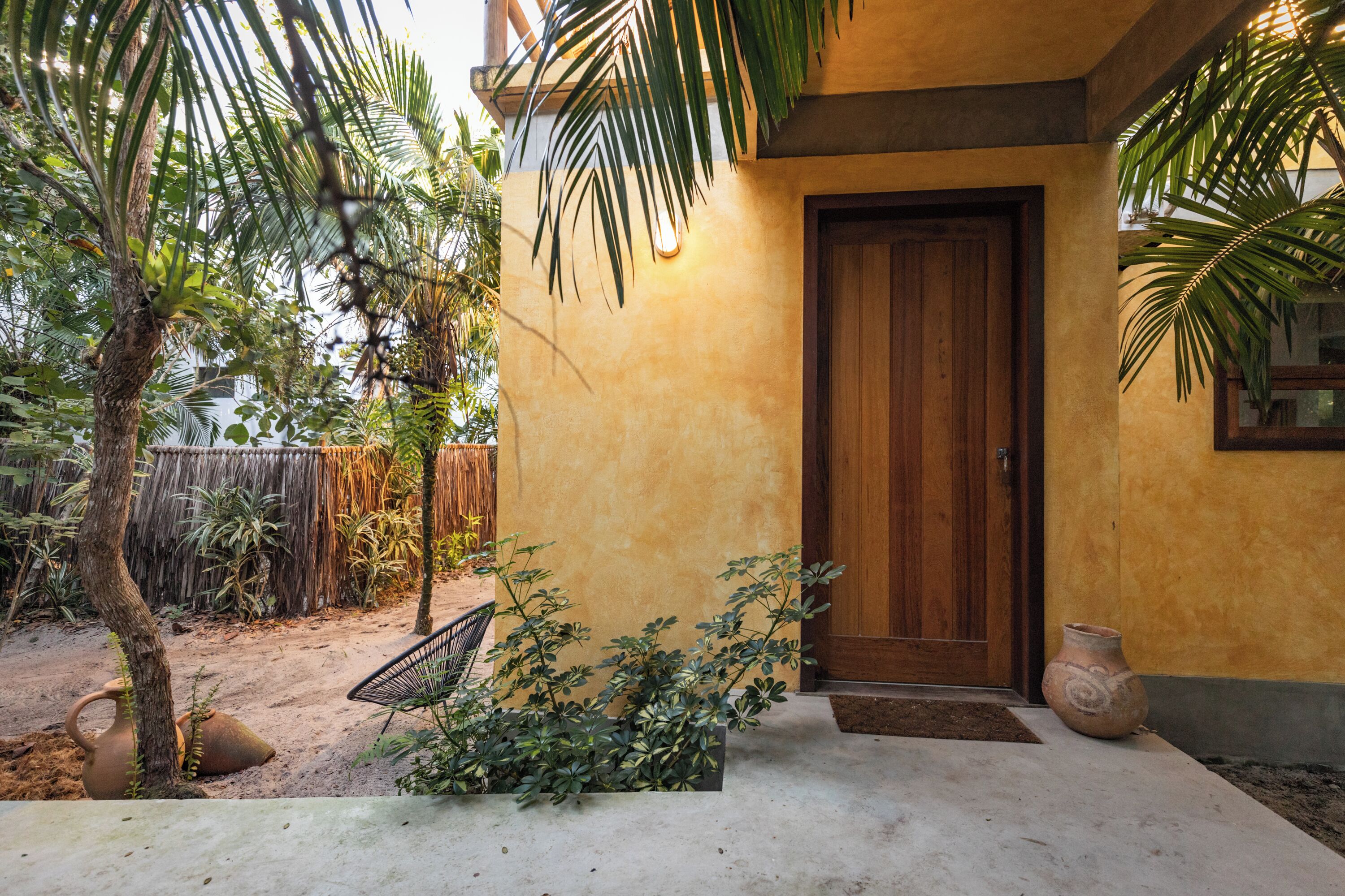 Una puerta de madera, un sendero de hormigón, una planta pequeña en maceta, una silla de mimbre y una maceta de barro.