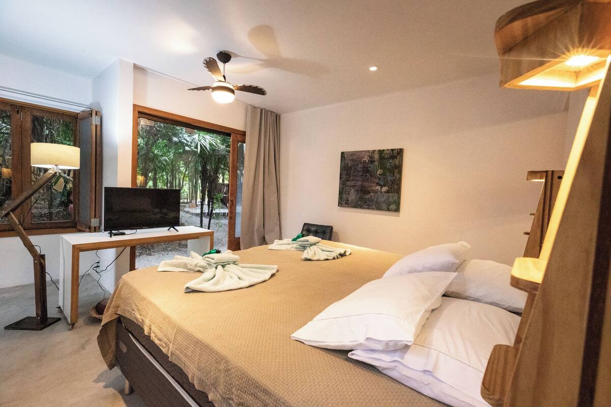 Una camera d'albergo con un letto, una scrivania con un computer, una ventola a soffitto e una finestra con tende.