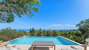 Une piscine rectangulaire, avec une table en bois et deux chaises longues au premier plan, entourée d’une végétation luxuriante et sous un ciel bleu clair.