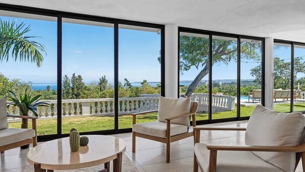 Un balcon spacieux avec de grandes fenêtres, une chaise en bois, une table ronde et une vue sur la mer et la verdure.