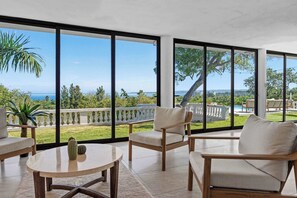 Un balcon spacieux avec de grandes fenêtres, une chaise en bois, une table ronde et une vue sur la mer et la verdure.