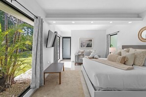 A modern bedroom with a large bed, a TV mounted on the wall, and a view of greenery outside. - Sun Bliss | Stunning Infinity Pool & Ocean Views (Aguada)