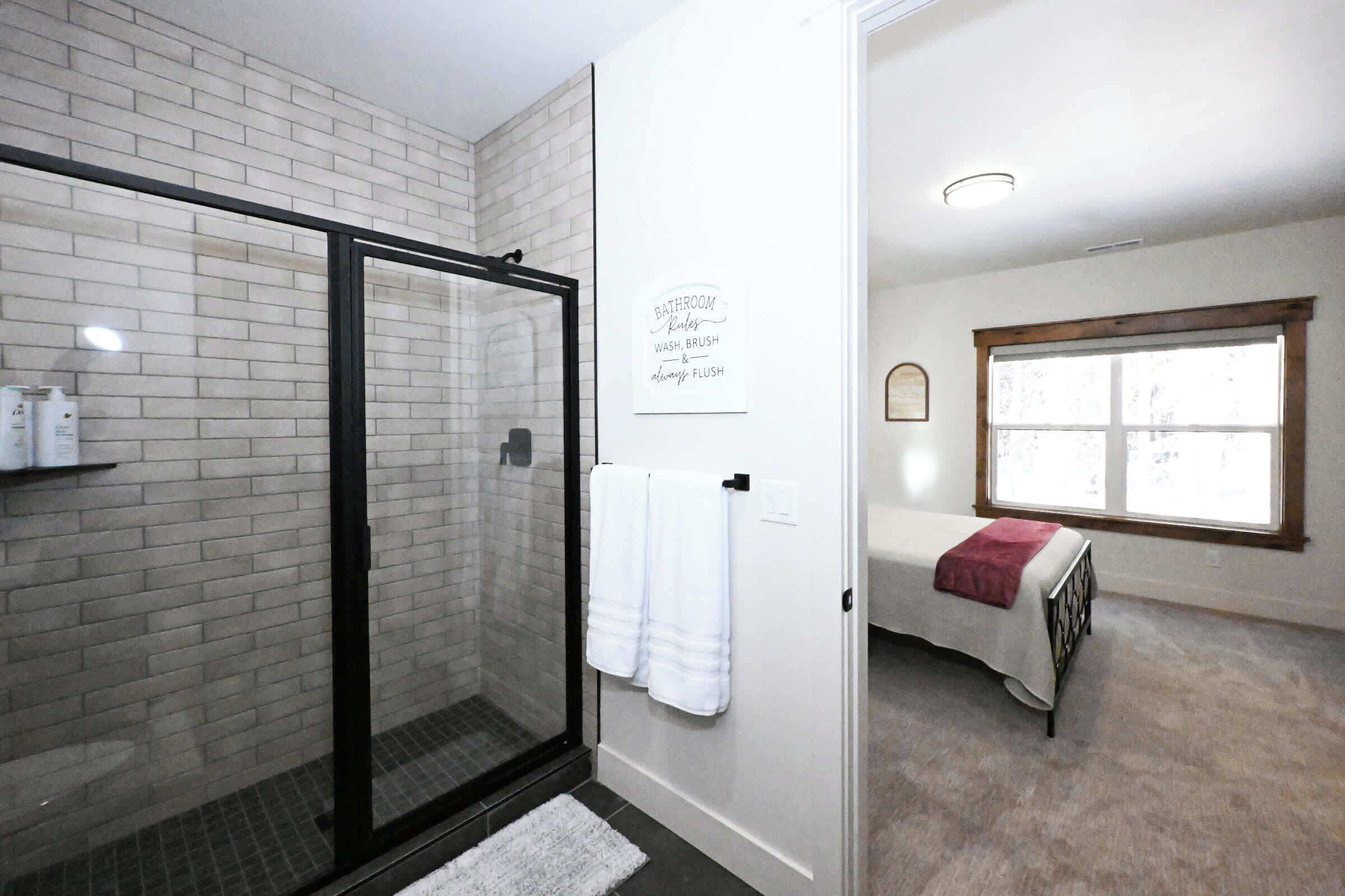 A bathroom with a white tiled wall, a glass shower enclosure, a white towel hanging on the door, and a window with a view.