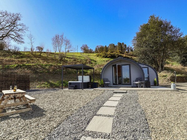 Una estructura moderna y parcialmente abierta con una zona de estar techada, una pequeña mesa con una botella, y un sendero de piedra que la conduce.