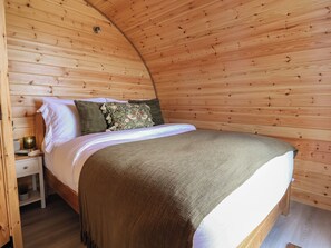 Una habitación acogedora de madera con techo curvo, una cama con manta verde, almohadas blancas y una mesita de noche con lámpara.