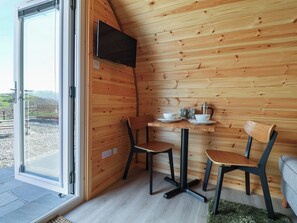 Interior de una cabaña de madera con televisión, dos sillones, una mesa con tazas y un hervidor, y una vista a través de una puerta de cristal.