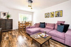 A living room with a purple sofa, wooden coffee table, and a dining area with a wooden table and chairs. - Open Mind Property - Hot Tub Retreat Sleeps 9 (Bournemouth)