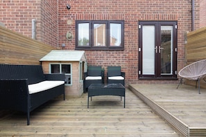 An outdoor patio with a brick wall, two windows, a sliding glass door, and outdoor furniture including a sofa, chairs, and a table.