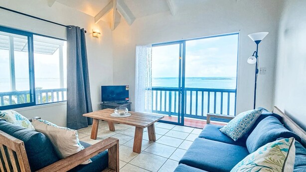 A living room with a sofa, a wooden coffee table, a TV, and a balcony with a view of the sea. - Toreaiva 91 Beachfront Studio - Upper (Arorangi)