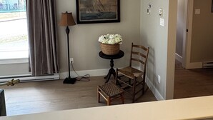 Une pièce avec vue sur l’extérieur, un tableau encadré, un panier en osier posé sur une petite table, une chaise et une lampe.