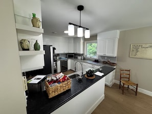 A modern kitchen with white cabinets, a black countertop, a microwave, a refrigerator, and a basket of snacks. - Getaway to Mahone Bay Nova Scotia (Mahone Bay)