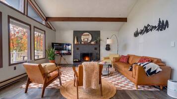 A living room with a fireplace, a sofa, a TV, and a large window with autumn leaves.