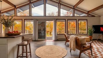 A spacious room with a high ceiling, large windows, a kitchen area, and a seating area with a woven rug.