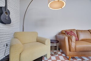 A room with a beige armchair, a brown leather sofa, a guitar on the wall, and a round table with a lamp. - Summer Oasis: Hot Tub, Huge Yard, Pool + Lakes (Long Pond)