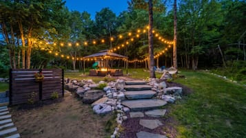 Un espacio de estar rústico al aire libre con una chimenea, rodeado por un sendero de piedra y una valla de madera.
